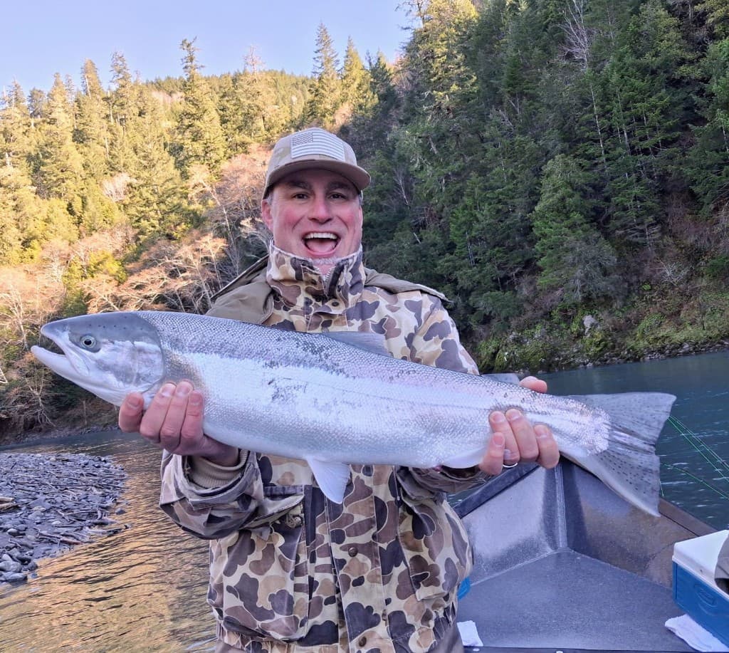 Grinning angler holding a large chrome steelhead in a forested river canyon